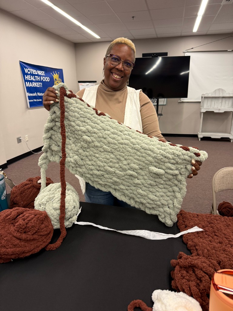 A smiling participant at the Fall into Cozy workshop holds up a sage green chunky-knit blanket behind a table of oversized yarn