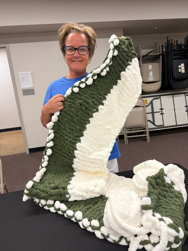 A smiling participant holding up a finished forest green and cream chunky hand-knit blanket with white pom-pom edges