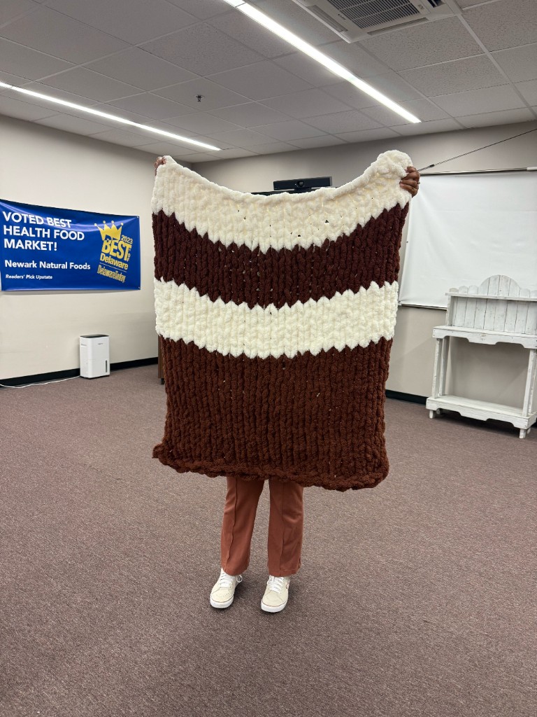 A participant holds up a finished chunky-knit blanket with dark brown and cream stripes at the Fall into Cozy workshop