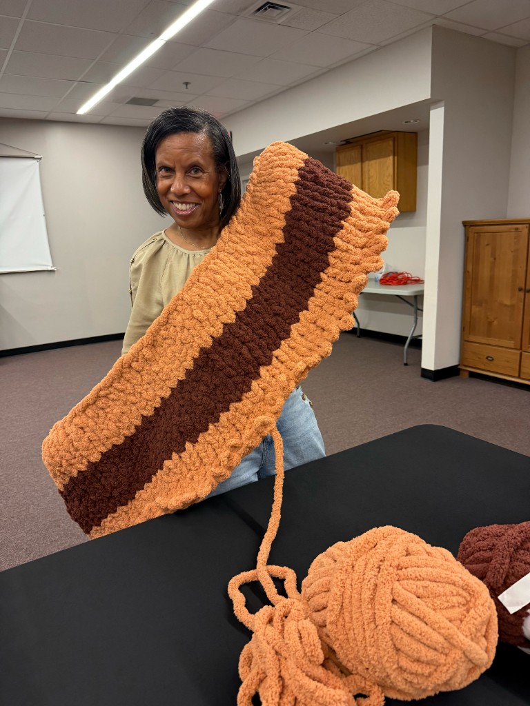 A participant proudly displays a burnt orange and dark brown striped chunky hand-knit blanket in progress