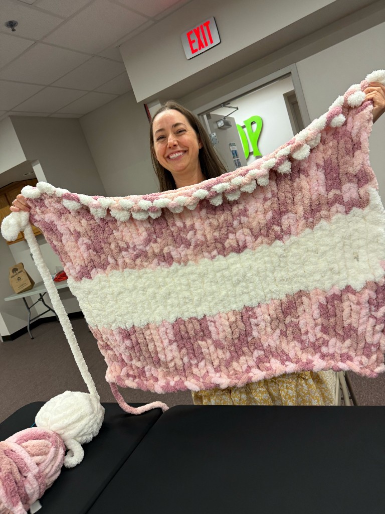 A smiling participant displays a large hand-knitted chunky yarn blanket with white and pink stripes