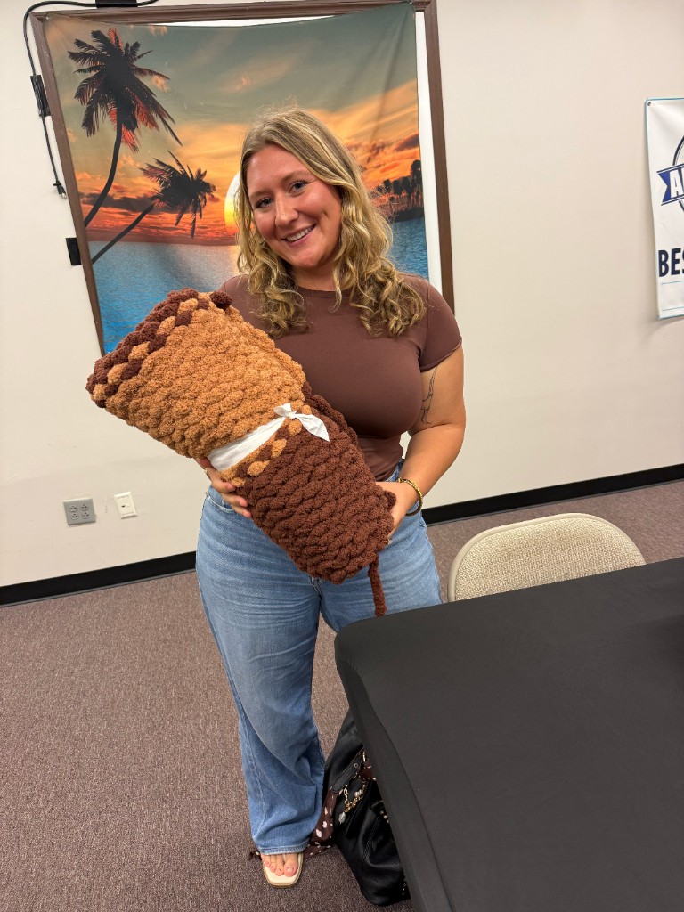 A smiling participant proudly displaying a completed brown and tan chunky knit blanket tied with a white ribbon
