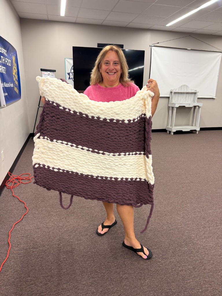 A smiling participant holding up a finished purple and cream striped chunky knit blanket at Wrapped in Strength