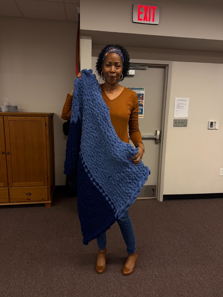 A participant holding a blue ombre chunky knit blanket in front of a workshop door