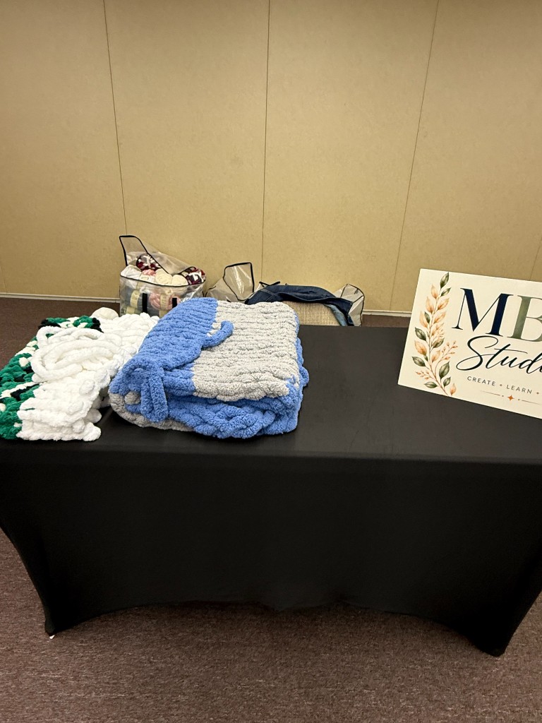 Finished blue, white, and green chunky blankets folded on a table beside the MBD Studio sign
